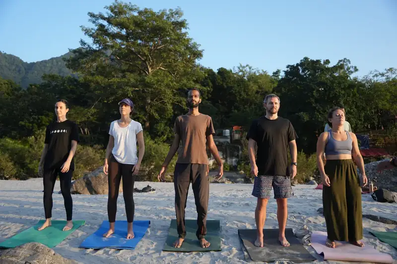 Yoga students standing in Tadasana on the Ganga riverbank at sunrise in Rishikesh